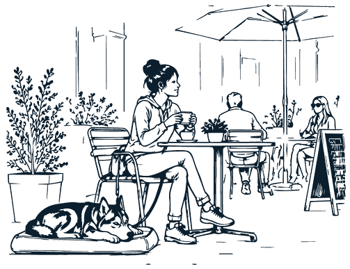 A dog lying on a mat under a café table while a woman drinks her coffee